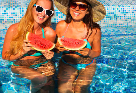 Two sexy women with dark hair eating watermelon in the poolの写真素材