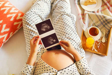 Pregnant woman wearing pijama with book, tea, cake relaxing at home. Top view の写真素材