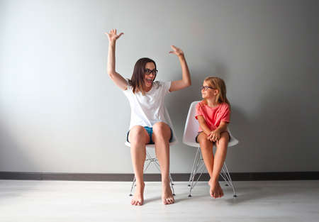 Happy mother and her little daughter sitting on the chairs by the grey wall. Portrait of mother and daughter with eyeglasses. Funny and happy motherの写真素材