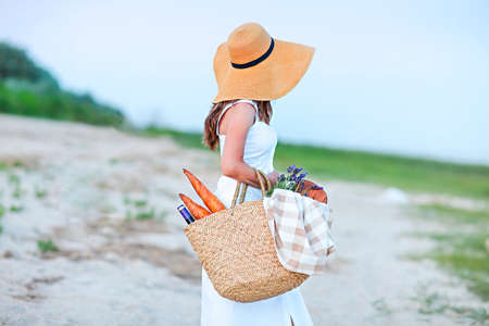 Young woman holding picnic basket with bottle of wine and baguette on sand beachの写真素材