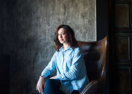 Young brunette woman sitting in armchair by the grey wall smiling at the cameraの写真素材