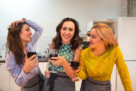 Cheerful female friends preparing a meal together and drinking red wine in the kitchen