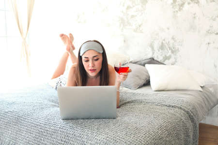Cheerful brunette woman holding glass of wine wearing sleaping mask in bed. Reading and looking at laptopの写真素材