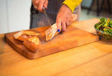 Cropped shot of woman cutting baguette bread on the tabletop at the kitchen