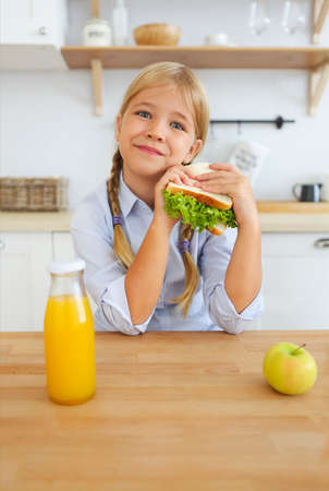 Happy little girl of school age, blonde kid enjoying healthy breakfast eating sandwich and fruits and drinking orange juice sitting at bright sunny kitchenの写真素材
