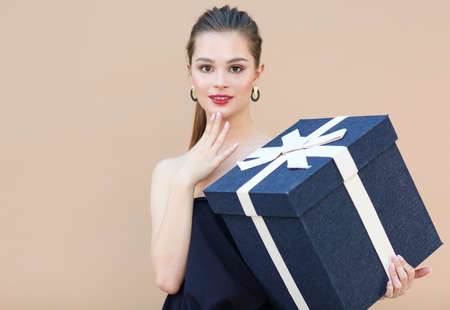 Surprised cute girl in navy blue clothes holding big gift box isolated on beige