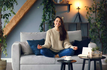 Body and spirit health. Cute smiling woman is learing yoga at home sitting on the sofaの写真素材