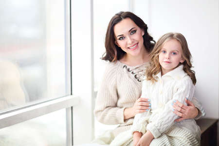 Happy loving family together. Mother and child daughter hugging by the window wearing cozy sweaters