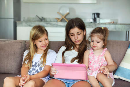Three kid sisters girls playing together with tablet pc on white sofa in the living roomの写真素材