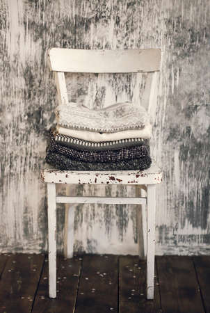Pile of knitted warm grey and white blankets, scarves and woolen sweaters for winter or fall cold weather on white stool near white and gray wall, copy space. Cosy home winter or autumn atmospereの写真素材