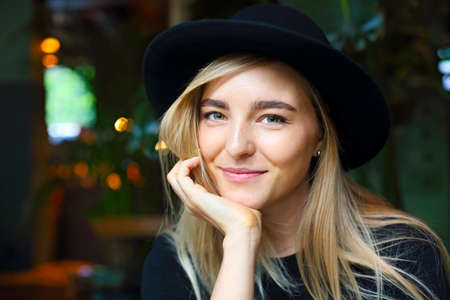 Attractive cute caucasian girl in black tshirt and black hat, sitting at cafe table by the window alone, looking outside with thoughtful expression and smilingの写真素材