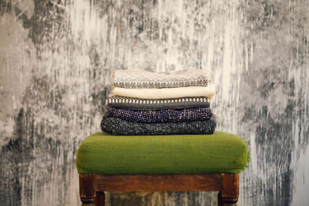 Pile of knitted warm grey and white blankets, scarves and woolen sweaters for winter or fall cold weather on white stool near white and gray wall, copy space. Cosy home winter or autumn atmospereの写真素材