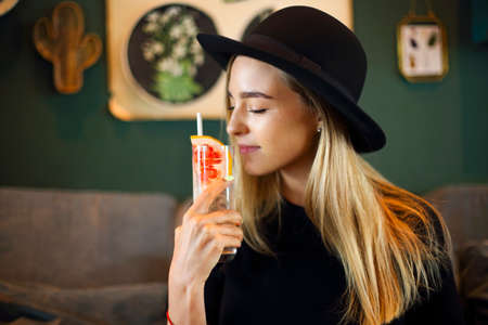 Attractive cute caucasian girl in black tshirt and black hat drinkig cold citrus juice or lemonade,...