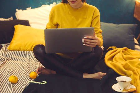 Beautiful young woman shopping online on laptop in cozy Christmas interior. Female sitting on the bed next the Christmas tree, bying gifts for the family on winter salesの写真素材