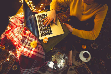 Beautiful young woman shopping online on laptop in cozy Christmas interior. Female sitting on the...