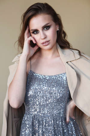 Beautiful young fashion model wearing a trench coat and sequins dress over beige backgroundの写真素材