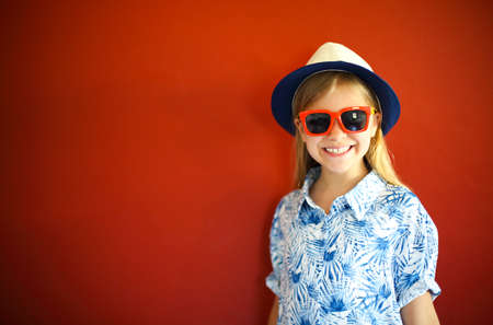 Pretty emothional child wear a hat and sunglasses on a red background. Copy space, daylight