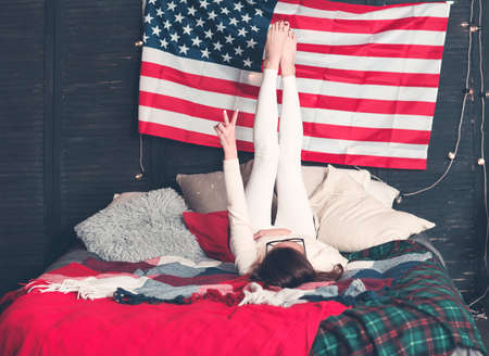 Pretty female in casualwear on bed with American flag on the wall during celebration of national Independence day の写真素材