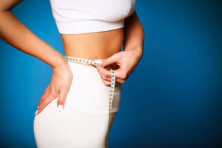 Beautiful skinny slim girl measuring her thin waist by tape measure at the blue backgriund. Sport and Healthy lifestyle concept の写真素材
