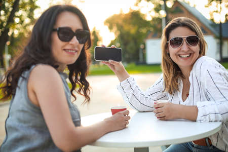 Smiling adult female friends sitting with coffee cups at table on street showing blank smartphone at camera の写真素材