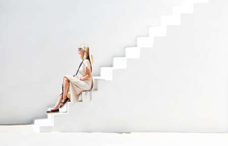 Side view of female in sundress and sunglasses sitting on plaster white stairway in sunshine