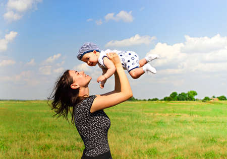 Side view of cheerful female holding adorable baby up standing against lush green fields under blue...