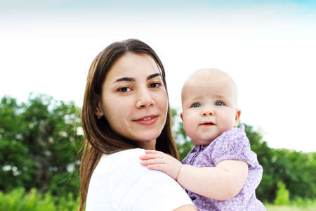 Cheerful female holding adorable baby up standing against lush green fields under blue skyの写真素材