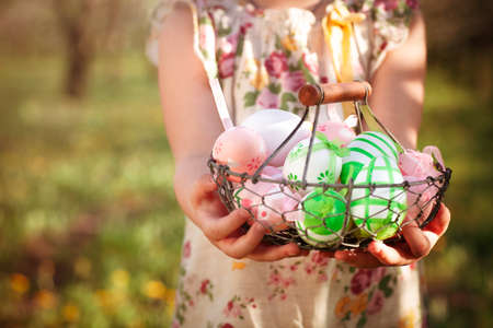 Crop little girl in flowered dress demonstrating to camera metal basket with pink and green painted...
