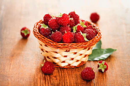Small wicker bowl with ripe red raspberry on wooden table with few berries and green leavesの写真素材