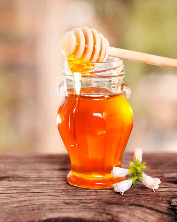 Stylish glass jar with golden honey and dipper on wooden table with flower on blurred backgroundの写真素材
