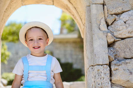 Happy little boy in light summer wear and straw hat smiling and looking at camera while standing in...