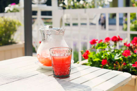 From above of glass with fresh red drink and jug with cold beverage on wooden white table in garden on sunny summer dayの写真素材