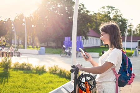 Teeen female in eyeglasses in a white clothes is using her smartphone app to remotely activate and prepay a modern rental push e-scooter or an e-bicycle outdoorsの写真素材