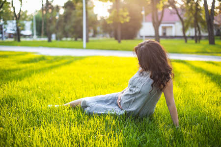 Anonymous pregnant female touching belly gently and sitting on green grass while relaxing in park during sunset in summerの写真素材