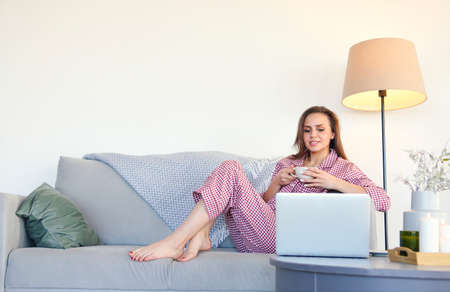 Barefoot woman in pajama enjoying hot beverage and watching film on laptop while resting on sofa in...
