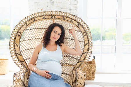 Dreamy pregnant woman in summer outfit looking away while resting in comfortable wicker armchair on...