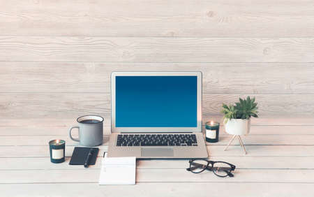Modern laptop with blue screen placed near mug of hot beverage and various decorations on wooden table in cozy home officeの写真素材