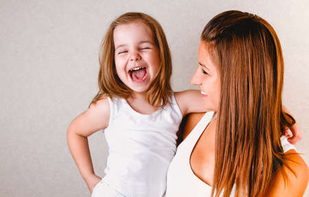 Smiling young casual redhead female holding laughing little daughter in white tank top while having fun together standing against light backgroundの写真素材