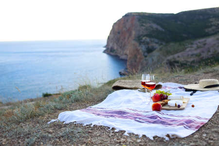 Set for romantic picnic with glasses of wine with fruits and cheese on blanket on green meadow at rocky seaside in summer eveningの写真素材