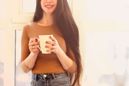 Young female in casual wear sitting on windowsill and drinking tea while resting at homeの写真素材