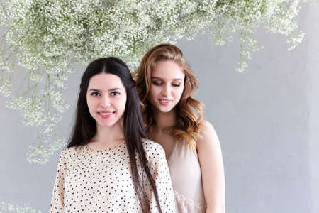 Two beautiful multicultural young women posing camera over grey background with delicate flowers