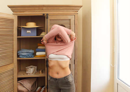Young girl takes off a stylish pink sweater by the cupbord in the room at homeの写真素材
