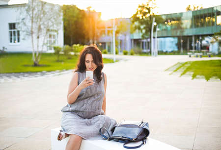 Happy pregnant woman holding cell phone while sitting on park bench outdoors