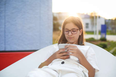 Beautiful young hipster teen girl in glasses using smart phone at bench in the park at sunset timeの写真素材