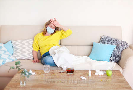 Ill mature lady in medical mask touching hot forehead while sitting on couch near table with various medicationsの写真素材