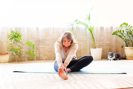 Full body adult female in casual clothes raising arms and stretching body while doing yoga on floor...