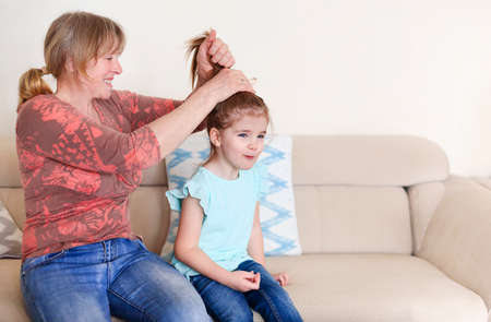 Happy mature woman smiling and doing ponytail to adorable little girl while sitting on sofa at home...