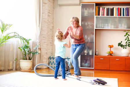 Full length happy mature woman with vacuum cleaner giving high five to little girl while tidying cozy living room at home togetherの写真素材