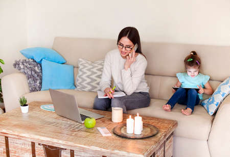 Positive young woman in casual clothes discussing business issue and taking notes in planner while working on remote project in cozy living room near little daughter playing video game on tabletの写真素材