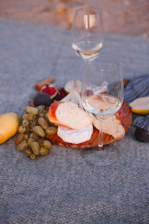 Top view of wooden board with pieces of bread pear figs nuts green grape and delicious cheese with white wine on blanketの写真素材